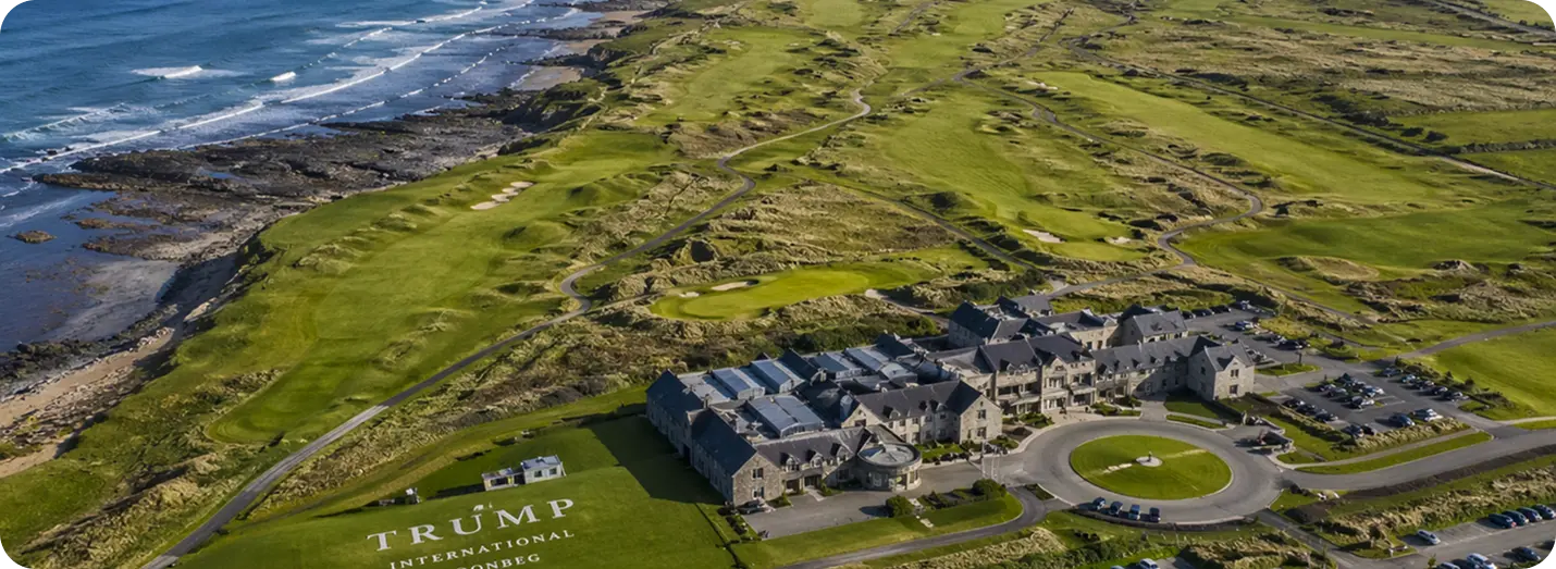 Aerial view of Trump International Golf Links Doonbeg, featuring the large stone hotel clubhouse building and expansive green fairways along a rugged, rocky coastline.