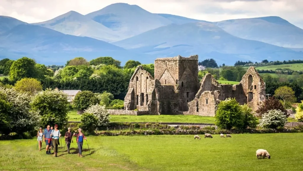 Hikers passing medieval abbey ruins in Ireland's countryside with mountains behind