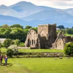 Hikers passing medieval abbey ruins in Ireland's countryside with mountains behind